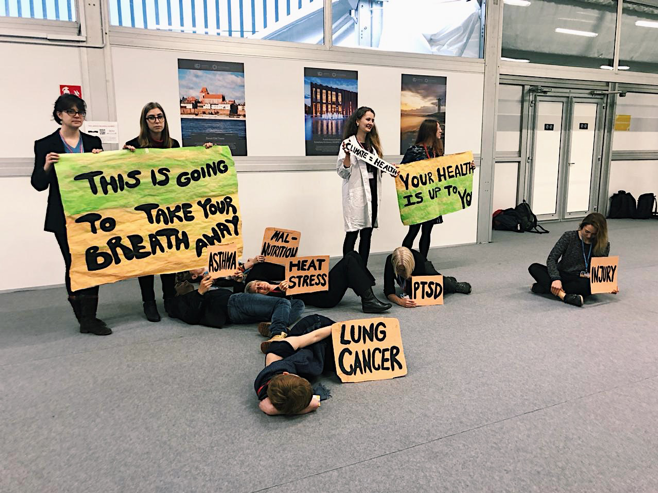 En grupp unga som står framför en vägg med bilder från Polen. Gänget håller målade plakat där det står “this is going to take your breath away”, “your health is up to you”, “PTSD”, “lung cancer” och “heat stress”. 
